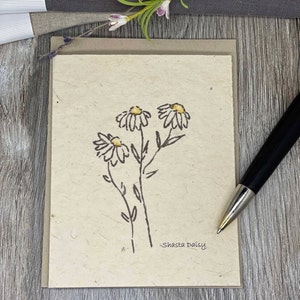 May include: A simple line drawing of three Shasta Daisies on a brown card with a textured surface. The flowers are drawn in black ink with yellow centers. The text "Shasta Daisy" is written in black ink at the bottom of the card.