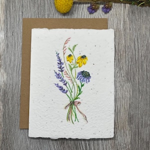 May include: A rectangular white greeting card with a textured, speckled surface featuring a watercolor illustration of a wildflower bouquet. The bouquet includes yellow and purple flowers tied with a thin brown ribbon at the base.