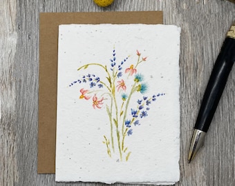 May include: A rectangular white card with a deckled edge features a watercolor illustration of a wildflower bouquet with blue, pink, and green stems. A brown envelope sits behind the card, and a black ballpoint pen rests to the right.