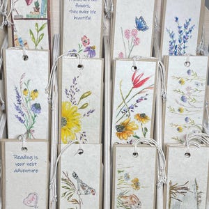 May include: A display of handmade paper bookmarks with floral and nature-themed illustrations. Some bookmarks feature text, including "Friends are like flowers, they make life beautiful" and "Reading is your next adventure."