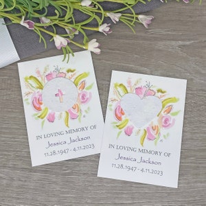 May include: Two white cards with watercolor floral designs. Each card has a small circle or heart-shaped cutout with a seed paper insert. The text on the cards reads "In loving memory of Jessica Jackson 11.28.1947 - 4.11.2023".