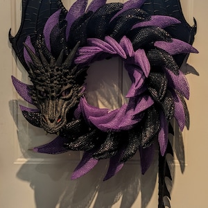 May include: A dragon-themed wreath with a black dragon head, wings, and tail. The wreath features a circular design with alternating layers of black and purple mesh. The dragon's head has red eyes and detailed features.