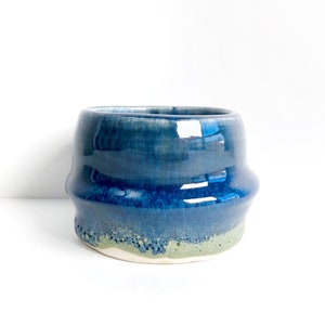May include: A small, handmade ceramic bowl with a blue and green glaze. The bowl has a rounded shape and a slightly uneven rim.