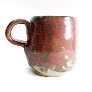 Puede incluir: Una taza de cerámica hecha a mano con un esmalte rojo y marrón. La taza tiene una forma única e irregular y una superficie ligeramente texturizada.