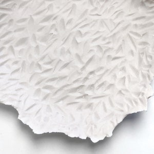 May include: White ceramic plate with a textured surface. The plate has a rough, uneven edge and a pattern of raised lines that resemble leaves.