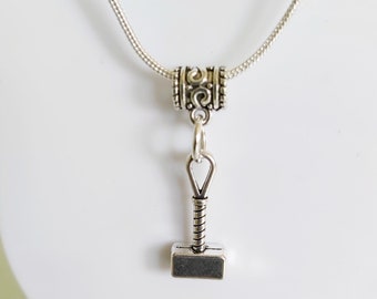 Op de afbeelding: Zilverkleurige ketting met een hanger in de vorm van Thors hamer, Mjolnir. De hanger heeft een rechthoekige kop en een opgerolde handgreep. De ketting wordt gepresenteerd op een witte buste.