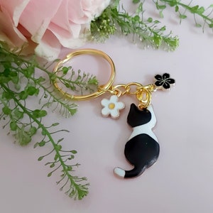 ゴールドトーンのスプリットキーチェーンに、白のデイジーと黒のサクラのチャームが付いた、黒と白の花柄の猫のキーリング。小さな花柄のかわいい猫。