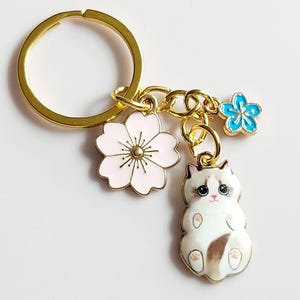 Könnte beinhalten: Goldfarbener Schlüsselanhänger mit Ring, Kette und drei Anhängern. Zu den Anhängern gehören eine rosa Blume, eine blaue Blume und eine weiß-braune Katze. Die Katze liegt auf dem Rücken. Der Schlüsselanhänger befindet sich auf weißem Hintergrund.
