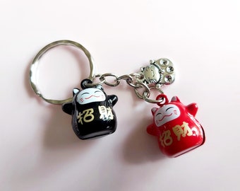May include: A black and red lucky cat keychain with a silver charm. The black cat has gold writing that says "招財" and the red cat has gold writing that says "招財".