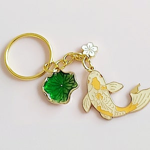 May include: Gold-tone keychain featuring a white and yellow koi fish charm, a green leaf charm, and a white flower charm. The keychain also includes a gold-tone ring.