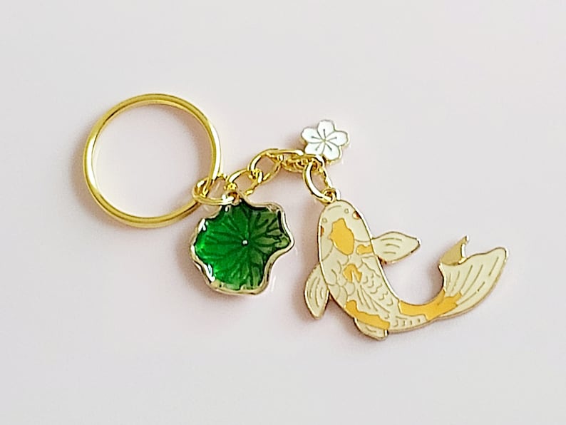 May include: Gold-tone keychain featuring a white and yellow koi fish charm, a green leaf charm, and a white flower charm. The keychain also includes a gold-tone ring.