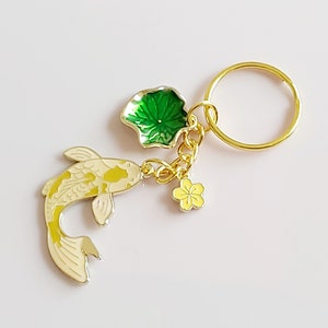 May include: Gold-tone keychain featuring a white and yellow koi fish charm, a green lily pad charm, and a yellow flower charm.
