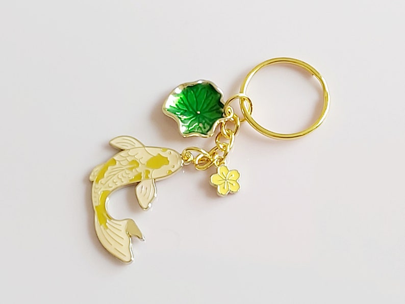 May include: Gold-tone keychain featuring a white and yellow koi fish charm, a green lily pad charm, and a yellow flower charm.