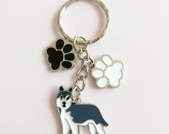 May include: A silver keychain with a husky dog charm and two paw print charms. The husky charm is black, white, and gray. One paw print is black, and the other is white. The keychain is on a pink background.