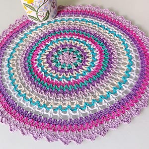 May include: A crocheted doily with a circular design in shades of purple, pink, white, and teal. The doily has a scalloped edge.