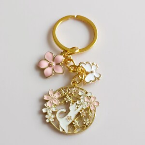 May include: Gold-coloured keychain with a floral and cat design. The keychain features a gold ring, pink and white flower charms, and a circular pendant with a white cat reaching for flowers. The pendant is surrounded by pink and white flowers.