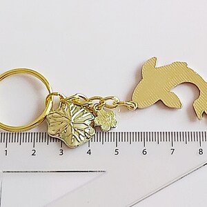 May include: Gold-colored keychain featuring a fish charm, a flower charm, and a small charm with a leaf design. The keychain is attached to a gold-colored key ring. The charms are hanging from a gold-colored chain.