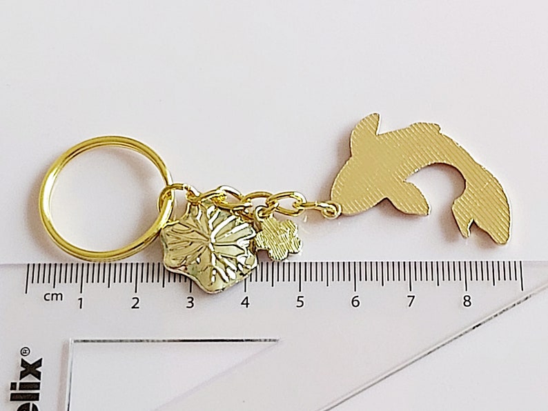 May include: Gold-colored keychain featuring a fish charm, a flower charm, and a small charm with a leaf design. The keychain is attached to a gold-colored key ring. The charms are hanging from a gold-colored chain.