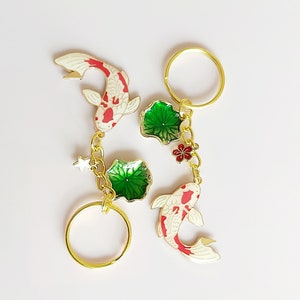 May include: Two gold keychains with red and white koi fish charms, green lotus leaf charms, and a small red flower charm. The keychains are attached to gold rings.