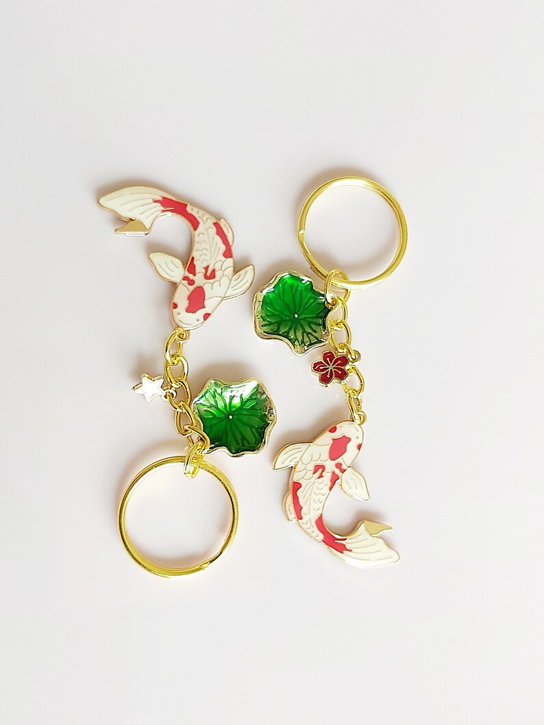 May include: Two gold keychains with red and white koi fish charms, green lotus leaf charms, and a small red flower charm. The keychains are attached to gold rings.