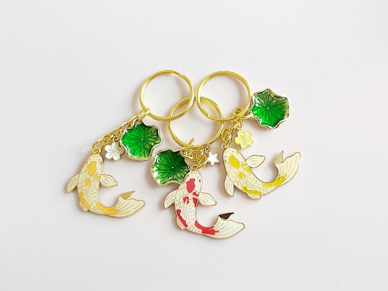 May include: Three gold keychains with charms of koi fish and green lily pads. The koi fish are white with yellow, red, and orange accents. The lily pads are green with gold accents.
