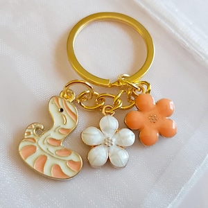 May include: Gold-coloured keyring with three charms: a snake in white and peach, a white flower, and an orange flower. The keyring is on a white, textured surface.