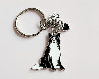 May include: A silver-colored keychain featuring a black and white dog charm. The dog charm is a sitting Border Collie with a white chest and face. The keychain also includes a flower and heart charm.
