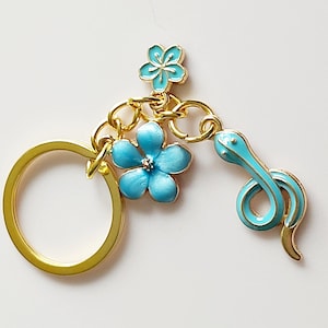 May include: A gold-coloured keyring featuring a turquoise snake charm and two turquoise flower charms. The snake and flowers have gold-coloured outlines. The keyring is set against a white background.