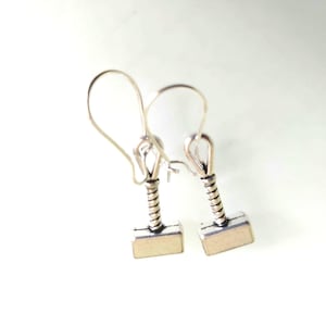 Pode incluir: Um par de brincos prateados em forma de martelo de Thor, Mjolnir. Cada brinco apresenta uma cabeça retangular e uma pega enrolada, com um gancho curvo para a orelha. Os brincos estão sobre um fundo branco.