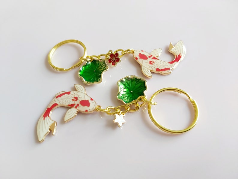 May include: Gold-tone keychain with two white and red koi fish charms, two green lotus leaf charms, and a gold star charm. The keychain also has two gold key rings.