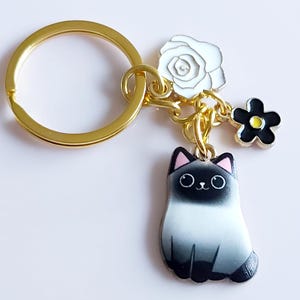Può includere: Portachiavi color oro con un ciondolo a forma di gatto siamese, una rosa bianca e un fiore nero con centro giallo. Il ciondolo a forma di gatto è nero, bianco e grigio. L'anello del portachiavi misura circa 3 cm di diametro.
