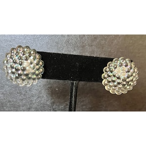 May include: A pair of clear, round stud earrings. Each earring is composed of numerous small, clear, spherical beads, creating a textured, clustered appearance. The earrings are displayed on a black stand against a dark background.