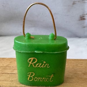 May include: A small, green plastic bucket with a gold-colored wire handle. The words "Rain Bonnet" are written in gold script on the front. The bucket is sitting on a wooden surface.
