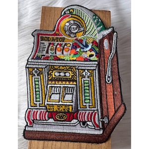 May include: Embroidered patch depicting a vintage slot machine. The patch features a colorful design with the text "ROL-A-TOP" and "JACK POT". The machine has a lever on the side and a coin slot. The patch is attached to a wooden surface.
