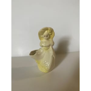 May include: A ceramic planter shaped like a girl in a yellow dress and hat. The planter has a small opening in the front for planting.