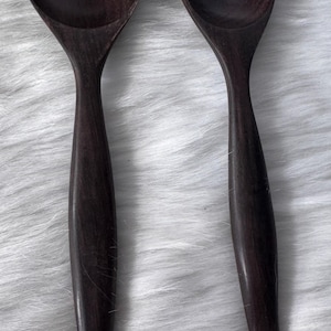 Puede incluir: Juego de cuchara y tenedor de madera marrón oscuro para servir ensalada. La cuchara tiene un cuenco redondeado y el tenedor tiene tres púas. Ambos utensilios tienen mangos largos y delgados. El juego es de madera oscura con veta visible.