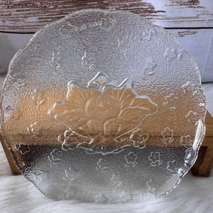 May include: Clear glass plate with a textured surface and a floral design. The plate features a raised flower in the center and smaller flowers around the edge. The plate is round and has a scalloped edge.