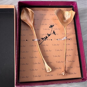 May include: A set of two gold-colored serving spoons in a decorative box. The spoons have a unique petal-like design and are presented on a brown background with the words "Table Art" printed repeatedly. The box has a pink border.