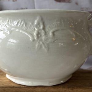 Peut inclure: Un pot de chambre en céramique couleur crème avec une finition brillante et une texture légèrement craquelée. L'extérieur est orné d'un motif floral en relief et comprend une anse incurvée sur le côté droit ainsi qu'un large bord évasé.
