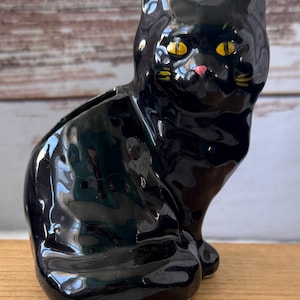May include: A glossy black ceramic cat figurine with yellow eyes and a pink nose. The cat is in a seated position with its ears perked up. The figurine is on a wooden surface.