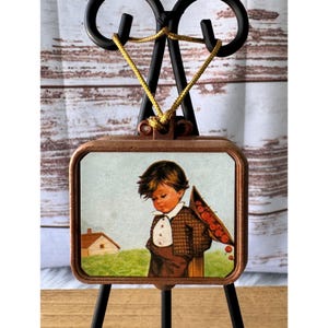 Puede incluir: Adorno enmarcado de estilo vintage con una impresión de un niño pequeño con una chaqueta marrón y un sombrero lleno de bayas rojas. El marco es de color cobre y el adorno cuelga de un cordón dorado. El adorno se exhibe en un caballete de metal negro.