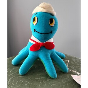 May include: A blue plush octopus wearing a white sailor hat, red bow tie, and a white and red striped collar.