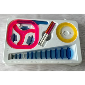 May include: A white plastic tray containing a selection of blue and pink plastic stencils for drawing circles, ovals, and other shapes. The tray also includes two white pens and a yellow stencil labelled "Ring Holder".