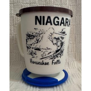 Könnte beinhalten: Eine weiße Keramiktasse mit braunem Deckel und einem blauen Untersetzer. Die Tasse zeigt eine Schwarzweißillustration der Niagarafälle und den Text "NIAGARA Horseshoe Falls".