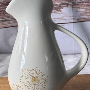 May include: Cream-colored ceramic pitcher with a curved handle and spout. The pitcher features two gold-colored dandelion designs on the front. The pitcher is likely used for serving beverages and has a glossy finish.