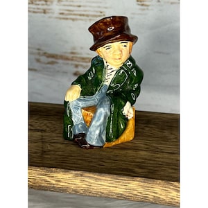 May include: A ceramic figurine of a man wearing a green coat, brown hat, and blue jeans. He is sitting on a small wooden stool.
