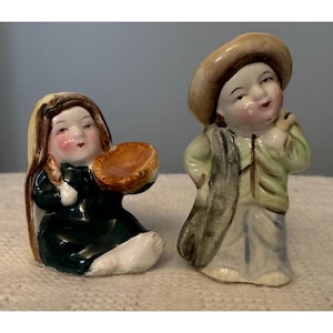 Puede incluir: Un par de saleros y pimenteros de cerámica vintage que representan a una niña sosteniendo una cesta y un niño con un sombrero y llevando un saco. La niña lleva un vestido verde y una cubierta de cabeza blanca. El niño lleva una camisa verde y pantalones azules. Los saleros están pintados a mano con detalles en marrón, verde, azul y blanco.