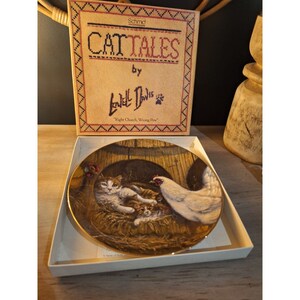 May include: A decorative plate featuring a cat and chicken scene, with the text "Cat Tales" by Lowell Davis. The plate is in a white box, with a cross-stitch design on the box cover.