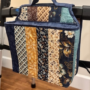 May include: A rectangular fabric storage bag featuring a patchwork design of vertical strips in navy blue, mustard yellow, cream, and brown with various floral and plaid patterns. The bag has a top handle and is attached to a metal mobility walker frame.