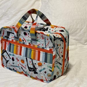 Peut inclure: Un sac de rangement matelassé rectangulaire avec une base en tissu blanc ornée d'un motif répétitif de machines à coudre, de ciseaux et de pelotes à épingles. Le sac comprend des fermetures éclair orange et une poignée rayée multicolore.